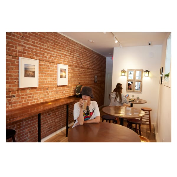 A woman in a bucket hat sips a drink at a round table in a cafe with brick walls.