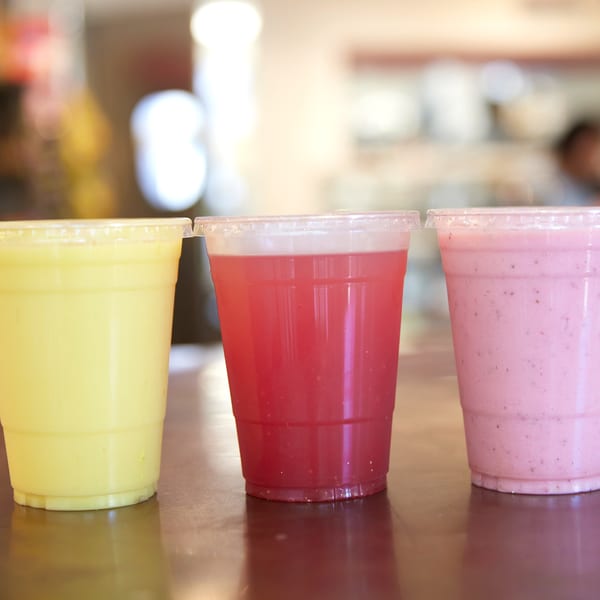 Three colorful drinks in plastic cups: yellow, red, and pink, on a brown surface.
