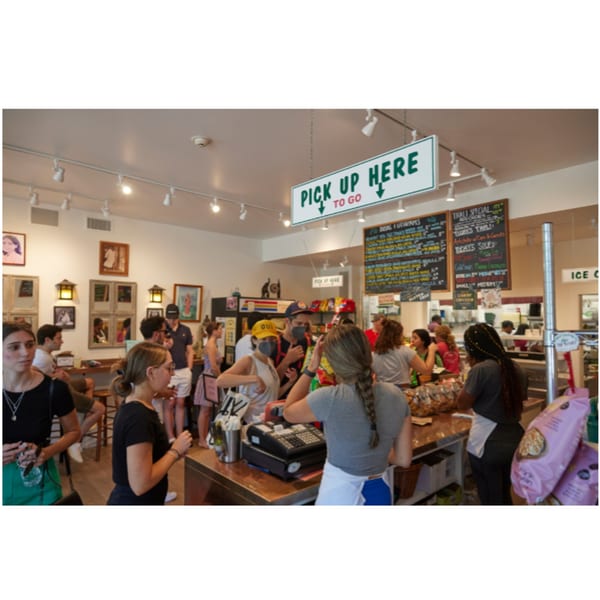 Customers and staff inside Hampton Chutney Co., with a "PICK UP HERE" sign and menu boards visible.