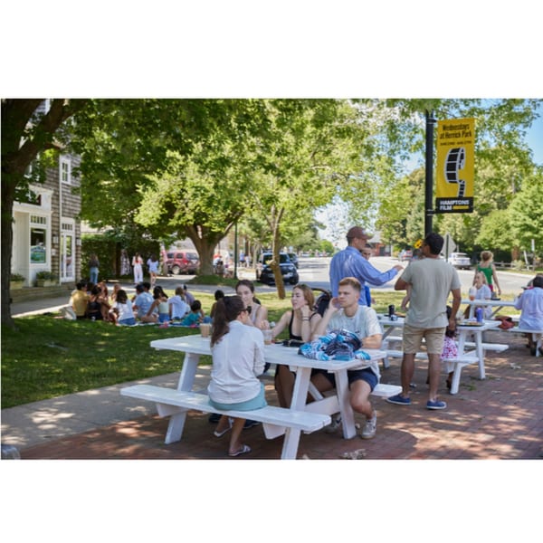 People enjoying the outdoor seating area with white picnic tables and green trees at Hampton Chutney Co.