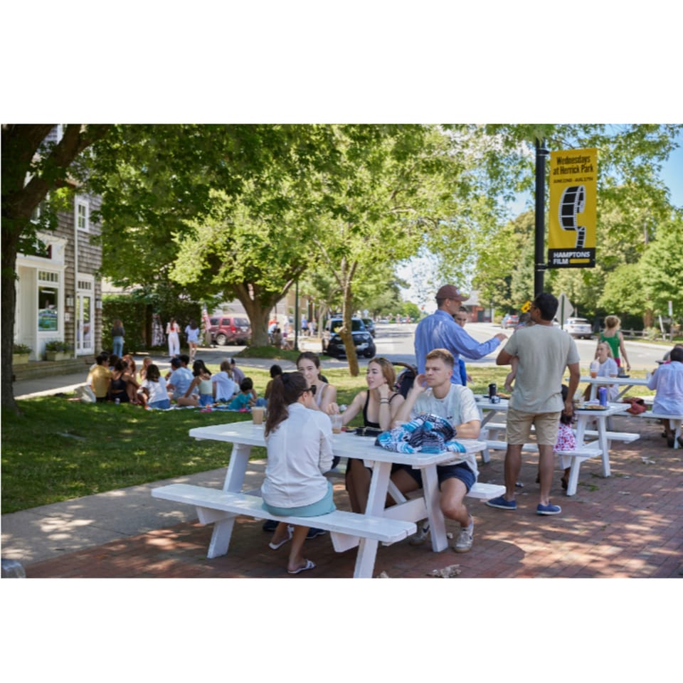 People enjoying the outdoor seating area with white picnic tables and green trees at Hampton Chutney Co.