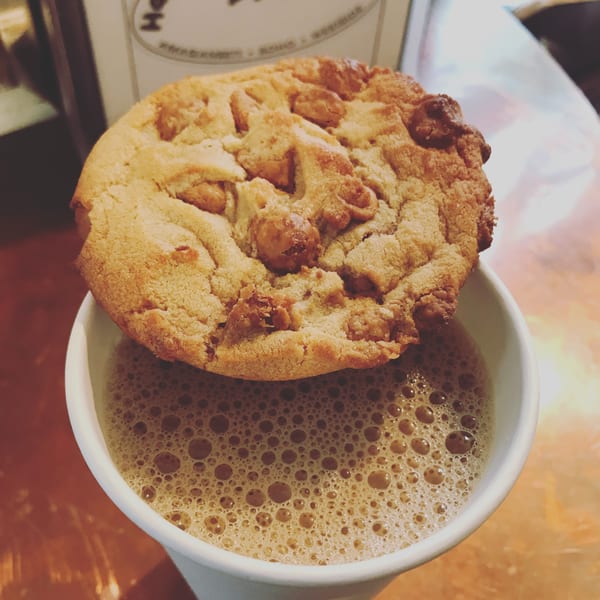 A large, golden cookie with nuts rests atop a white cup of frothy chai.