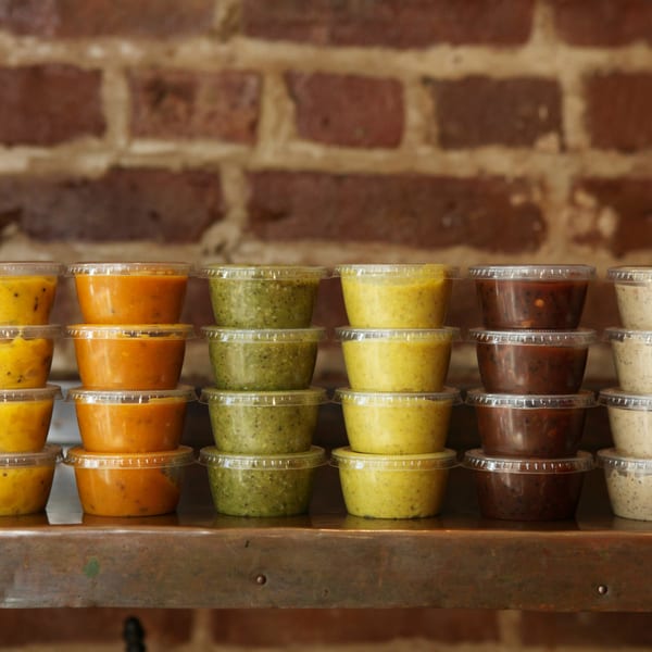 Rows of colorful chutneys in clear plastic containers, stacked on a metal shelf against a brick wall.