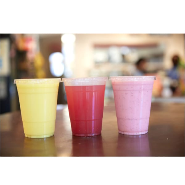 Three colorful drinks, yellow, red, and pink, in clear plastic cups on a counter.