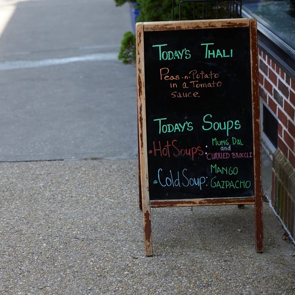 A Hampton Chutney Co. chalkboard sign on a sidewalk lists today's thali and soup specials.