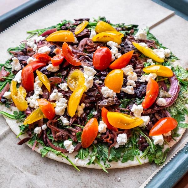 Unbaked Indian pizza with red onions, cherry tomatoes, feta, and greens on parchment paper.