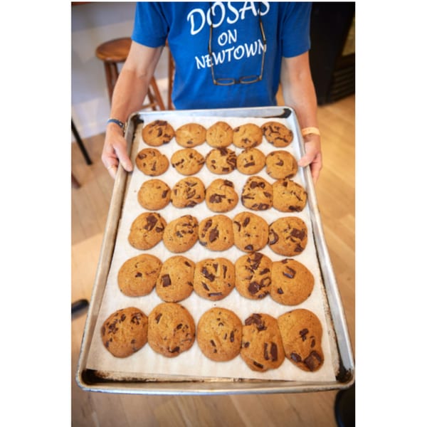 A person in a blue "DOSAS ON NEWTOWN" t-shirt holds a baking sheet filled with chocolate chip cookies.