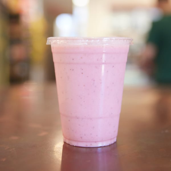 A pink lassi drink in a clear plastic cup with a lid, on a wooden table.