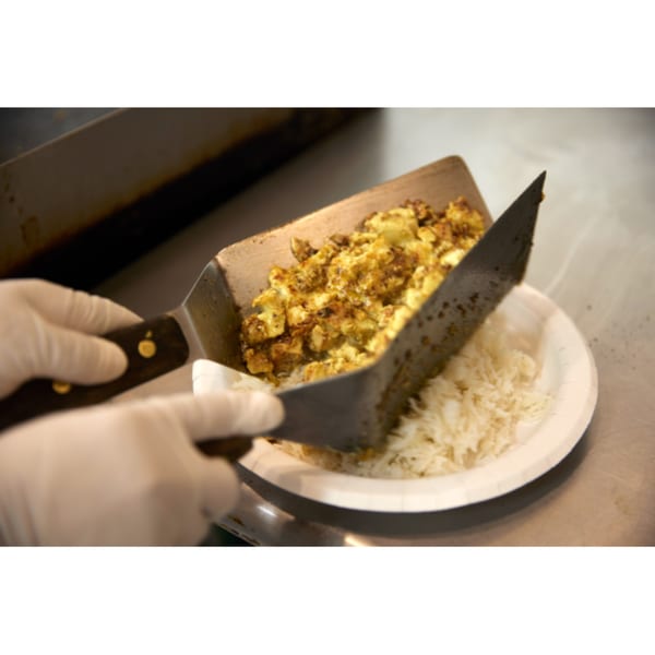 Gloved hands use spatulas to serve Indian spiced potatoes and rice onto a white plate.