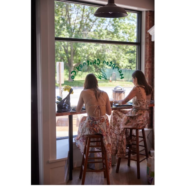Two women in floral dresses sit at a window bar inside Hampton Chutney Co., looking out at trees.